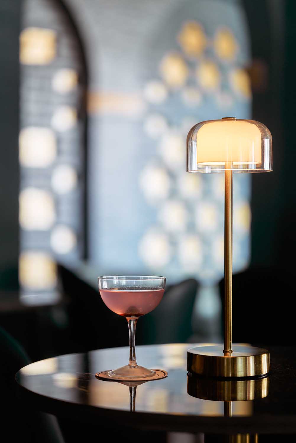 a rose-red cocktail in an up glass beside a modern lamp against a dark background